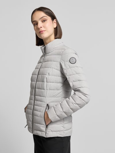 Modelka nosi kurtkę pikowaną z zamkiem błyskawicznym i kieszeniami zapinanymi na zamek błyskawiczny Steppjacke mit Reißverschlusstaschen Modell 'GS 111' od G.I.G.A. DX, krój slim fit, stójka, długość tyłu w rozmiarze 36: 67 cm, szerokość ramion w rozmiarze 36: 38 cm, szerokość klatki piersiowej w rozmiarze 36: 49 cm, długość rękawów w rozmiarze 36: 65 cm; jasnoszary kolor, widoczna tekstura pikowania, prosta forma; widok całej sylwetki z przodu