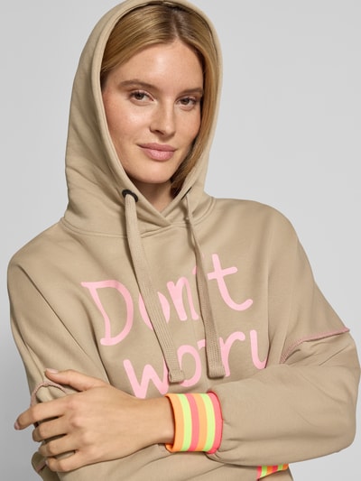 Frauenmodel trägt Oversized Hoodie mit Statement-Print von miss goodlife aus reiner Baumwolle in Beige mit Neon-Details, Nahaufnahme Oberkörper.