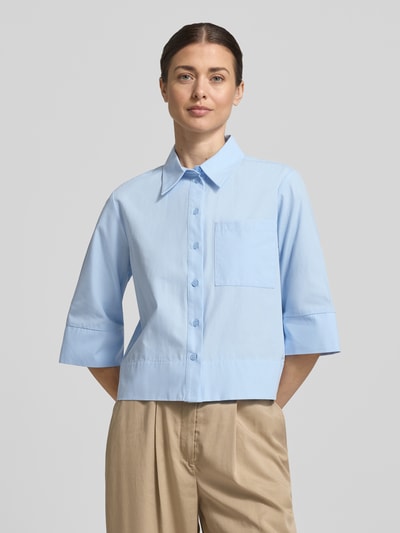 Modelka nosi Boxy Fit Bluse mit Brusttasche Modell 'Zarba Bold' od Someday, jasnoniebieska bawełna, kołnierzyk koszulowy, kieszeń na piersi, długość tyłu 54 cm, długość rękawa 39 cm, widok całej sylwetki z przodu