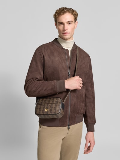Männermodel trägt Umhängetasche mit Logo-Print von Lacoste in Braun mit Muster, Herren Umhängetasche, Ganzkörperansicht von vorne.
