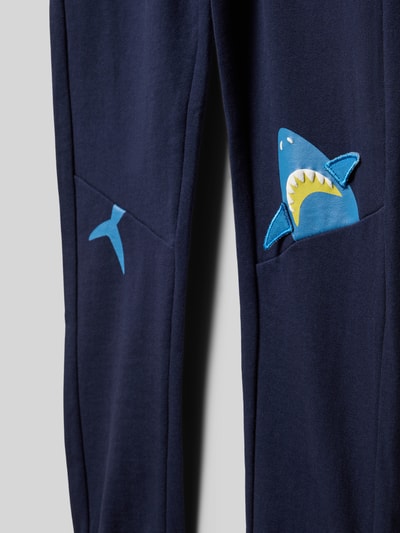 Produktfoto, Regular Fit Sweatpants mit Motiv-Print von Blue Seven in Blau, Reine Baumwolle, Eingrifftaschen, elastischer Bund mit Tunnelzug, elastische Beinabschlüsse, Innenbeinlänge 53 cm bei Größe 116, weiche Textur, sichtbar Hai-Motiv-Print auf der Tasche.