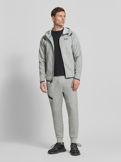 Männermodel trägt Regular Fit Jacke mit Kapuze Modell 'Unstoppable' von Under Armour in Hellgrau mit Logo, Zweiwege-Reißverschluss, Ganzkörperansicht von vorne. Elastischer Material-Mix mit Baumwoll-Anteil, geteilte Känguru-Tasche, elastische Abschlüsse.