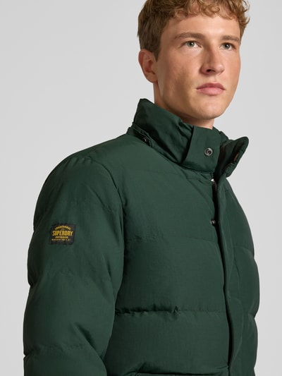 Männermodel trägt Jacke mit Kapuze Modell 'CLASSIC EVEREST' von Superdry in Dunkelgrün, sichtbarer Label-Patch, Nahaufnahme Oberkörper. Gerader Schnitt, Reiß- und Druckknopfverschluss, Kapuze mit Kordelzug, seitliche Eingrifftaschen, Ärmelriegel, Schulterbreite 47 cm, Rückenlänge 75 cm, Brustweite 58 cm, Ärmellänge 71 cm.