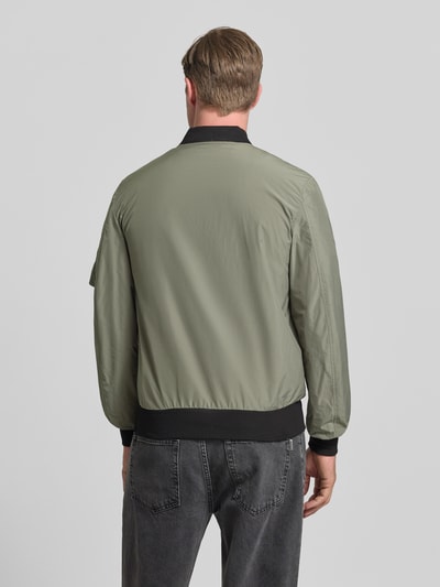 Männermodel trägt Bomberjacke mit Label-Patch und Eingrifftaschen von Original Bombers in Oliv mit Gerippte-Abschlüsse, Rückenansicht, Ganzkörperansicht Rückansicht. Herren Bomberjacke, Gerader-Schnitt, Reißverschluss, Reißverschlusstasche am Ärmel, Druckknopfverschluss, Rückenlänge bei Größe M: 67 cm, Schulterbreite: 43 cm, Brustweite: 53 cm, Ärmellänge: 68 cm.