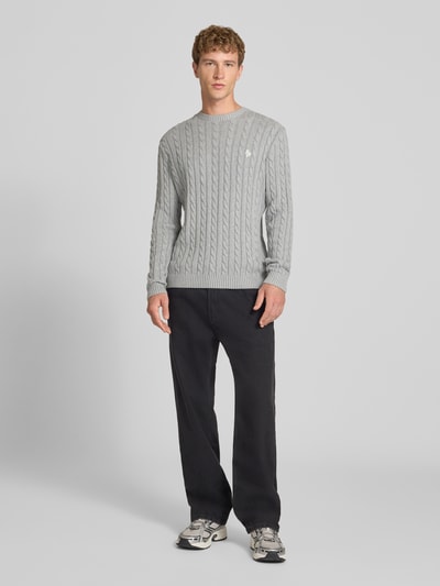 Männermodel trägt Slim Fit Strickpullover reine Baumwolle von REVIEW, graues Zopfmuster, gesticktes Logo, gerippte Bündchen, Ganzkörperansicht von vorne.