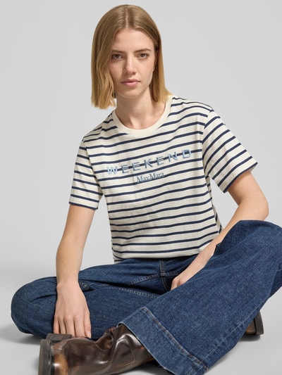 Frauenmodel trägt Relaxed Fit T-Shirt aus reiner Baumwolle Modell 'BORA' von Weekend Max Mara, cremefarben mit blauen Streifen und Motiv Stickerei, Jersey-Material, kurzarm, Rundhalsausschnitt, Nahaufnahme Oberkörper.