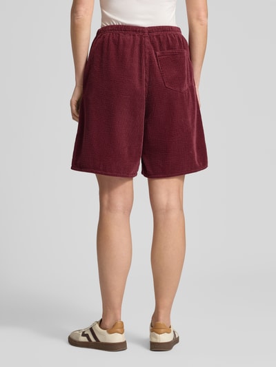 Männermodel trägt Shorts mit elastischem Bund Modell 'PADOW' von American Vintage, bordowa wiskoza, luźny krój, kieszeń z tyłu; Szerokość w rozmiarze S: 37 cm, długość nogawki wewnętrznej: 20 cm; Ganzkörperansicht Rückansicht.