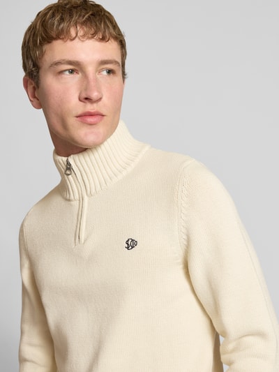 Männermodel trägt Slim Fit Pullover in Strick-Optik von Superdry, cremefarben, Stehkragen, Reißverschluss, Logo-Stitching, Nahaufnahme Oberkörper.