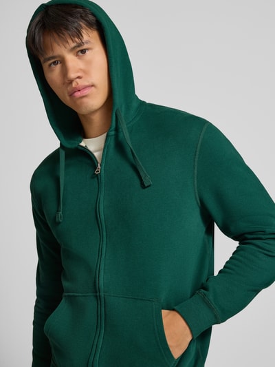 Männermodel trägt Regular Fit Sweatjacke aus Baumwoll-Mix mit Label-Detail von Superdry in Dunkelgrün, Nahaufnahme Oberkörper. Baumwoll-Mix, elastische Abschlüsse, Kapuze mit Tunnelzug, Känguru-Tasche, Rückenlänge 65 cm, Ärmellänge 65 cm.