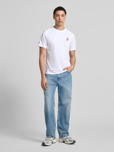 Männermodel trägt T-Shirt mit Logo- und Motiv-Print Modell 'SHORELINE' von Helly Hansen, weiße Reine Baumwolle, Gerader-Schnitt, Rundhalsausschnitt, Rückenlänge bei Größe M 70 cm, Ganzkörperansicht von vorne.