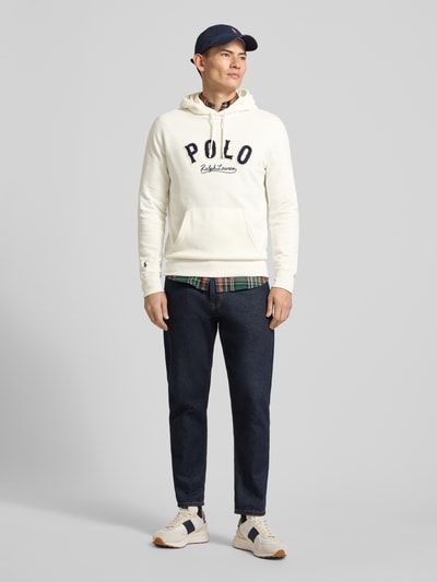 Männermodel trägt Hoodie mit Label-Stitching und Känguru-Tasche von Polo Ralph Lauren, cremefarben, kombiniert mit Jeans, Ganzkörperansicht von vorne. Baumwoll-Mix, Regular Fit, Rücken- und Ärmellänge passend zu Größe M.