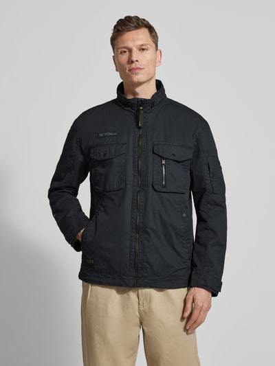 Męski model nosi Jacke aus reiner Baumwolle mit Brusttaschen od camel active, czarny kolor, prosty krój, stójka, naszywka z logo, widok całej sylwetki z przodu. Długość tyłu w rozmiarze 50: 73 cm, szerokość ramion w rozmiarze 50: 52 cm, szerokość klatki piersiowej w rozmiarze 50: 58 cm, długość rękawów w rozmiarze 50: 64 cm, zamki błyskawiczne, kieszenie na piersi i wpuszczane, patki przy mankietach, ochrona podbródka.