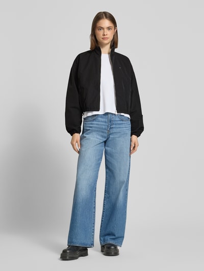 Modelka nosi Relaxed Fit Blouson mit Taillenbändern od Tommy Jeans, efekt: jednolity kolor, krój relaxed fit, stójka, zapięcie: zamek błyskawiczny, wyhaftowane logo, tasiemka w talii, długi rękaw, zakończenie rękawów: elastyczne, wpuszczane kieszenie z zapięciem na zatrzask, widoczna tekstura materiału, Ganzkörperansicht von vorne.