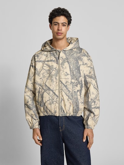 Männermodel trägt Jacke mit Kapuze und Logo von KARL KANI, Reine Baumwolle, Regular-Fit, Allover-Look, Beige-Grau, Ganzkörperansicht von vorne.