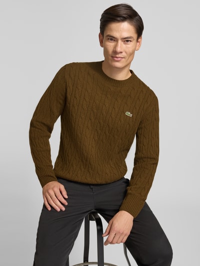 Männermodel trägt braunen Regular Fit Pullover aus reiner Wolle von Lacoste, Zopfmuster, neutrale Textur, Nahaufnahme Oberkörper. Herren Pullover von Lacoste.