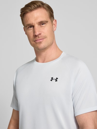 Männermodel trägt T-Shirt mit Label-Print Modell 'Tech' von Under Armour in Hellgrau, Rundhalsausschnitt, Gerader-Schnitt, Nahaufnahme Oberkörper. Herren T-Shirt, Rückenlänge bei Größe M: 75 cm.