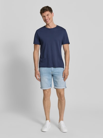 Männermodel trägt Slim Fit Jeansshorts aus Baumwoll-Mix Modell 'NAAILO' von Armedangels in Denim-Optik mit Label-Patch, 5-Pocket-Design, Beinabschluss mit Umschlag, Knopf und verdecktem Reißverschluss, Ganzkörperansicht von vorne.