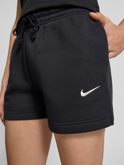 Frauenmodel trägt Sweatshorts mit Logo Modell 'Phoenix' von Nike aus schwarzem Baumwoll-Mix, Regular-Fit, elastischer Bund, Tunnelzug, Nahaufnahme Oberkörper. Innenbeinlänge 10 cm, Bundweite 37 cm bei Größe S, Logo seitlich sichtbar.