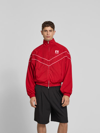 Männermodel trägt Bomberjacke mit Raglanärmeln von RHUDE in Rot, Zweiwege-Reißverschluss, Reißverschlusstaschen, lockere Passform, Ganzkörperansicht von vorne. Rückenlänge 61 cm, Schulterbreite 49 cm, Ärmellänge 67 cm, Größe M.