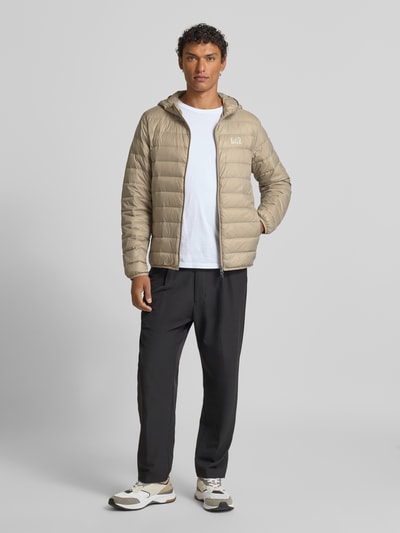 Męski model nosi Kurtka puchowa z zamkiem błyskawicznym od EA7 Emporio Armani, krój regular fit, kaptur, nadruk z logo, kieszenie z wszytym zamkiem błyskawicznym, długość tyłu w rozmiarze M: 68 cm, szerokość ramion w rozmiarze M: 44 cm, szerokość w rozmiarze M: 52 cm, długość rękawów w rozmiarze M: 61 cm, jasnobeżowe i czarne elementy, gładka tekstura, pełen widok z przodu.