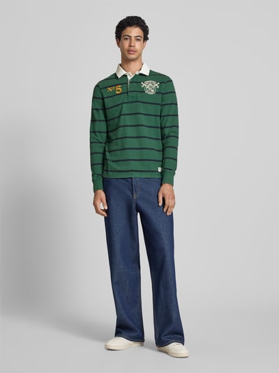 Männermodel trägt Regular Fit Rugby Shirt mit Logo- und Motiv-Stitching von Superdry in Grün mit Streifen und Label-Patch, Reine Baumwolle, Polokragen, kurze Knopfleiste, Rückenlänge bei Größe M: 70 cm, Ganzkörperansicht von vorne.