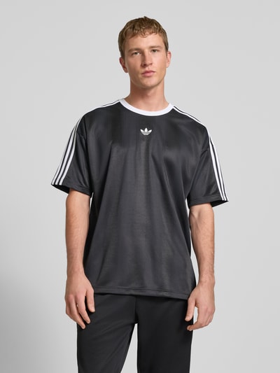 Männermodel trägt Funktionsshirt mit Logo und Rundhalsausschnitt von adidas Originals, schwarzes Shirt mit weißen Kontraststreifen, Ganzkörperansicht von vorne.