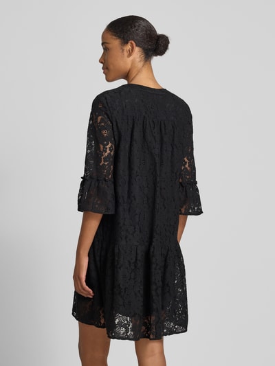 Frauenmodel trägt Spitzenkleid von Vero Moda, Modell 'HONEY', Optik: Unifarben, Passform: Loose-Fit, Länge: Knielang, Materialverarbeitung: Spitze, Baumwoll-Mix, Ärmelabschluss: Glockenärmel, Muster: Spitzen-Muster, Farbe: Schwarz, Ganzkörperansicht Rückansicht.