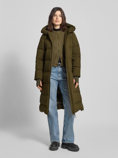 Modelka nosi Regular Fit Daunenmantel mit langen Storm Cuffs od Marc O'Polo w oliwkowej zieleni, krój regular fit, steppowana struktura, długi rękaw z Storm Cuffs, kaptur, dwukierunkowy zamek, kieszenie na suwak, z napoleońską kieszenią, wewnętrzne paski na ramię, materiał pikowany; widok całej sylwetki z przodu.