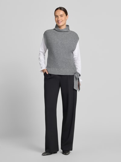 Frauenmodel trägt Relaxed Fit Pullunder aus reinem Kaschmir mit Stehkragen von Christian Berg Woman in Grau mit Zierschleife am Saum, ärmellos, kombiniert mit weißem Langarmshirt und schwarzer Hose, Ganzkörperansicht von vorne.