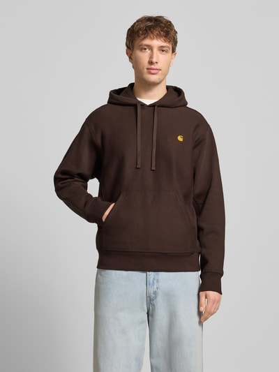 Männermodel trägt Hoodie mit Kapuze von Carhartt Work In Progress in Dunkelbraun, Baumwoll-Mix, Lockerer-Schnitt, Känguru-Tasche, Label-Stitching, gerippte-Abschlüsse, Tunnelzug, Ganzkörperansicht von vorne.