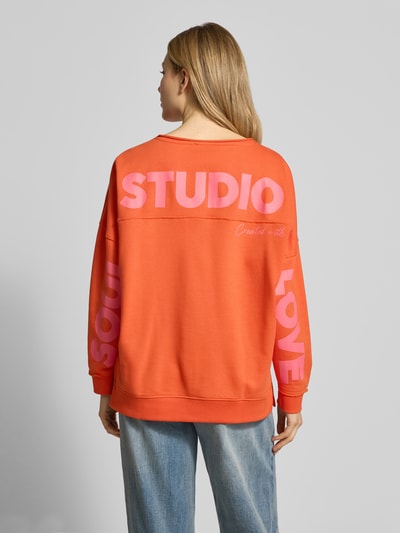 Modelka damska ma na sobie Oversized Sweatshirt mit Label-Print od Smith and Soul, bawełniano-modalowy, pomarańczowy z różowym nadrukiem, długi rękaw, ściągacze, widok całej sylwetki z tyłu.