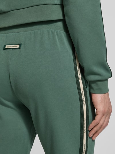 Männermodel trägt grüne Sweatpants aus Baumwoll-Mix von EA7 Emporio Armani mit seitlichem Logo-Streifen, Eingrifftaschen, Detailaufnahme.