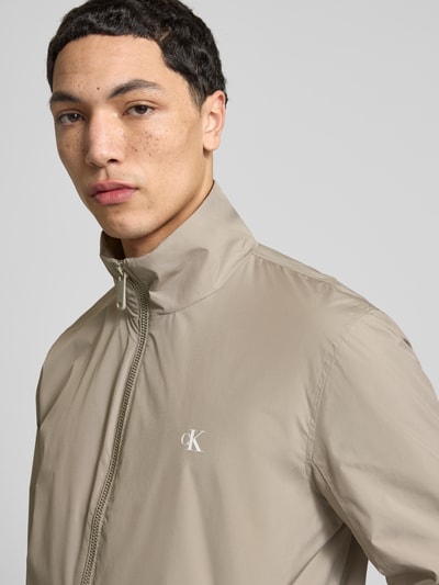 Mannelijk model draagt 'Trainingsjacke mit Logo und Stehkragen' van Calvin Klein Jeans, regular fit, logo, rits, ritszakken, beige, Nahaufnahme bovenlichaam.