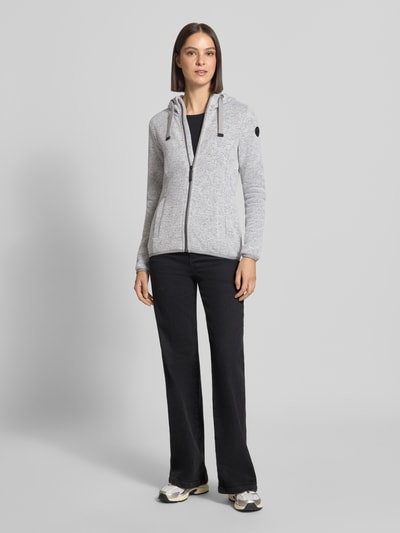 Frauenmodel trägt Regular Fit Sweatjacke mit Logo-Detail von G.I.G.A. DX in Hellgrau, Kapuze mit Tunnelzug, Reißverschluss, Eingrifftaschen, Strukturstrick, schwarze Hose, Sneakers, Ganzkörperansicht von vorne.