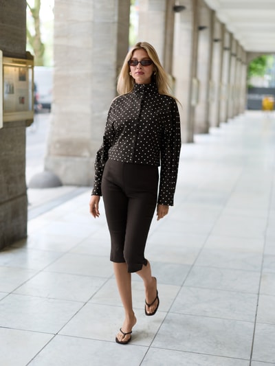 Frauenmodel trägt Kurze voluminöse Bluse - curated by Mary Braun von Style Icon Collection mit weißen Punkten, hohe Stehkragen, Baumwolle, Regular Fit, Knopfleiste, kombiniert mit knielanger Hose, Farben Schwarz und Weiß, glatte Stoffstruktur, Ganzkörperansicht von vorne.