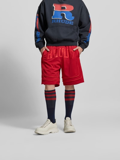 Männermodel trägt Baggy Fit Sweatshorts mit Tunnelzug von RHUDE aus rotem Mesh mit Label-Stitching, elastischem Bund, Eingrifftaschen, Nahaufnahme Oberkörper bis Knie.