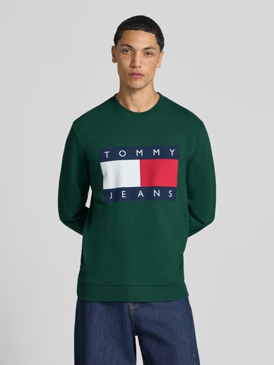 Männermodel trägt Regular Fit Sweatshirt aus reiner Baumwolle von Tommy Jeans, dunkelgrün mit buntem Logo, Ganzkörperansicht von vorne. Herren Sweatshirt von Tommy Jeans