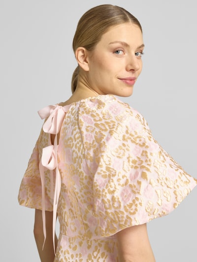 Vrouwenmodel draagt Tailliertes Cocktailkleid mit Allover-Muster Modell 'NORA' van Vero Moda met striksluiting, pofmouwen, rugdecolleté, all-over motief, getailleerd, minilengte, ronde hals, in roze en goud, detailopname rugdecolleté.