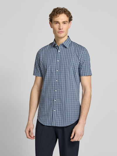 Męski model nosi Slim Fit Business-Hemd aus Baumwolle mit Kentkragen od Jake*s, niebieska krata, krój slim fit, czysta bawełna, krótki rękaw, guziki, widok całej sylwetki z przodu. Szerokość w rozmiarze 39/40: 48 cm, długość z tyłu: 79 cm, długość rękawa: 23 cm.