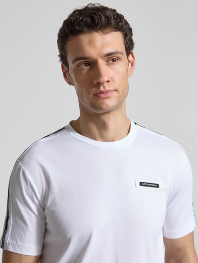 Mężczyzna model nosi Regular Fit T-Shirt mit Logo-Patch von EA7 Emporio Armani, biała bawełna, okrągły dekolt, zbliżenie na górną część ciała.