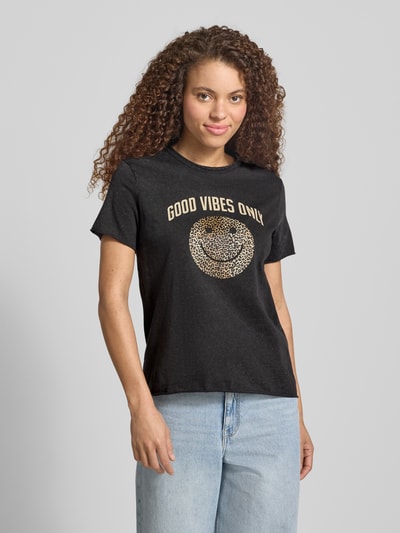 Frauenmodel trägt schwarzes Regular Fit T-Shirt aus reiner Baumwolle Modell 'LISA LIFE LUCY' von Only mit goldfarbenem Motiv-Print, Rundhalsausschnitt, kurzen Ärmeln und glatter Jersey-Oberfläche, Ganzkörperansicht von vorne.