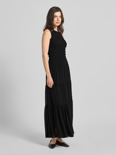 Frauenmodel trägt Maxikleid mit Smok-Details und Volantsaum von MICHAEL Michael Kors in Schwarz, Taillierter Schnitt, Rundhalsausschnitt, Rückenlänge bei Größe S: 141 cm, Brustweite bei Größe S: 31 cm, Ganzkörperansicht von vorne.
