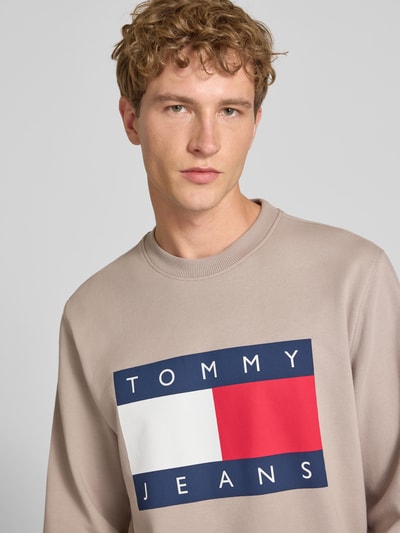 Mężczyzna nosi Regular Fit Sweatshirt aus reiner Baumwolle von Tommy Jeans, beżowa bluza z dużym logo na piersi, zbliżenie górnej części ciała.