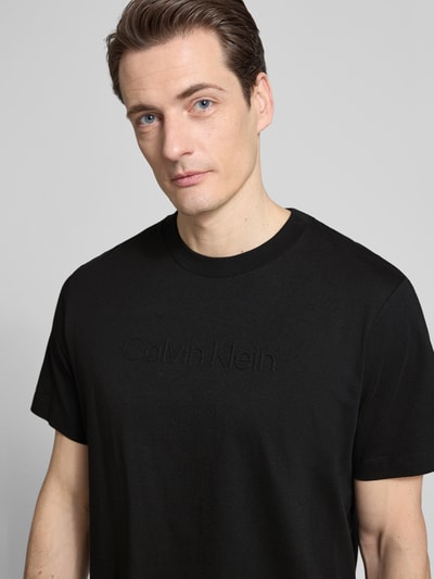 Männermodel trägt T-Shirt mit Rundhalsausschnitt von CK Calvin Klein, schwarze Farbe, Reine Baumwolle, Regular-Fit, Logo, Nahaufnahme Oberkörper. Rückenlänge bei Größe M: 67 cm.