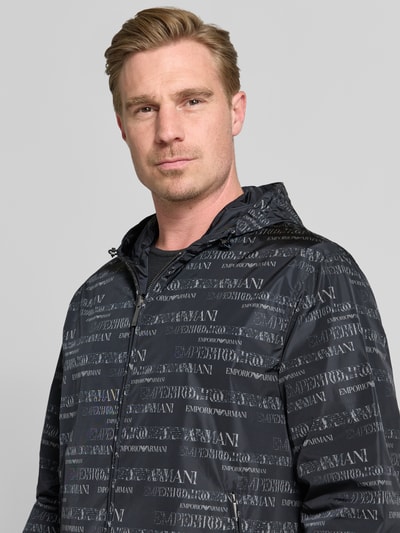 Männermodel trägt Jacke mit Logo-Muster und Zweiwege-Reißverschluss von Emporio Armani in Schwarz, Regular-Fit, Nahaufnahme Oberkörper. Rückenlänge 70 cm, Schulterbreite 58 cm, Brustweite 62 cm, Ärmellänge 66 cm, Kapuze mit Tunnelzug, Reißverschlusstaschen.