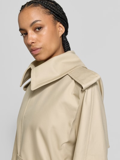 Frauenmodel trägt Trenchcoat aus reiner Baumwolle von Victoria Beckham in Beige, matter Stoff, Umlegekragen, Nahaufnahme Oberkörper, taillierte Passform, Pattentaschen.