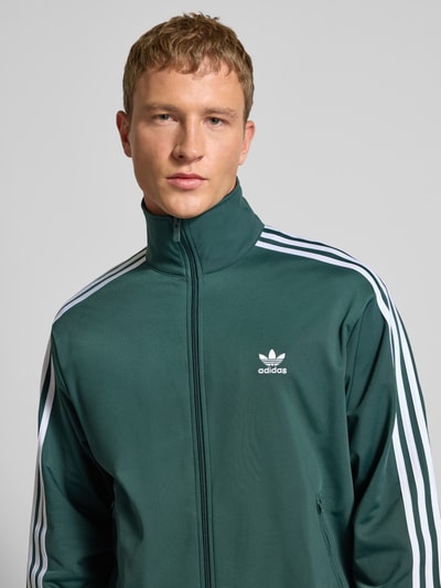 Männermodel draagt Regular Fit Sweatjacke mit Stehkragen Modell 'Bird' van adidas Originals, groene elastische materiaalmix, gestikt logo, steekzakken met ritssluiting, elastische boorden, Nahaufnahme Oberkörper.