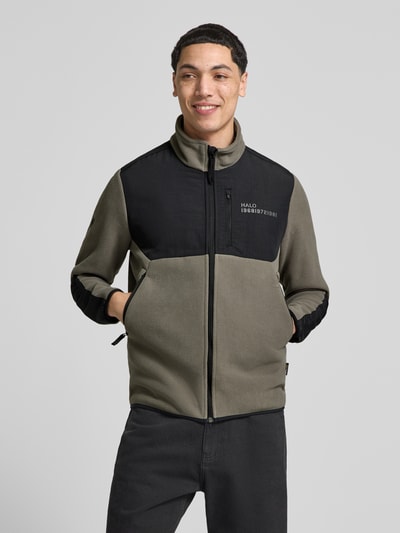 Männermodel trägt Fleecejacke mit Stehkragen und Reißverschlusstaschen von HALO in Olive und Schwarz, Label-Print, Regular-Fit, Ganzkörperansicht von vorne. Rückenlänge 71 cm, Schulterbreite 45 cm, Brustweite 45 cm, Ärmellänge 69 cm.
