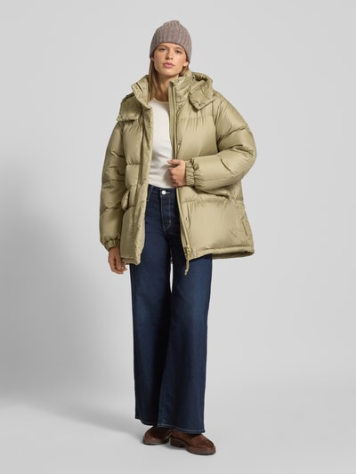Kobiece model nosi Oversized Daunenjacke mit abnehmbarer Kapuze od Marc O'Polo Denim, jasnobeżowy, pikowana tekstura, długie rękawy, widok całej sylwetki z przodu.