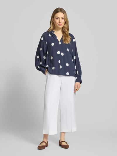 Frauenmodel trägt Bluse aus Viskose-Mix mit Serafino-Ausschnitt von Christian Berg Woman, dunkelblau mit weißen Punkten, Loose-Fit, Rückseite länger geschnitten, ganzkörperansicht von vorne.